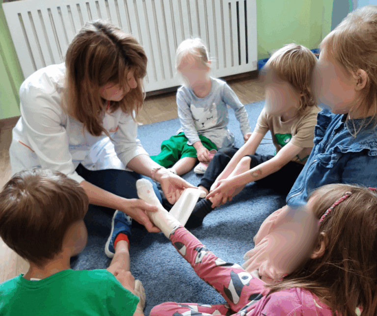 Warsztaty prozdrowotne dla przedszkoli w Warszawie – farmaceutka pokazuje dzieciom jak korzystać z bandaża, w jaki sposób owinąć prawidłowo stłuczoną kostkę.
